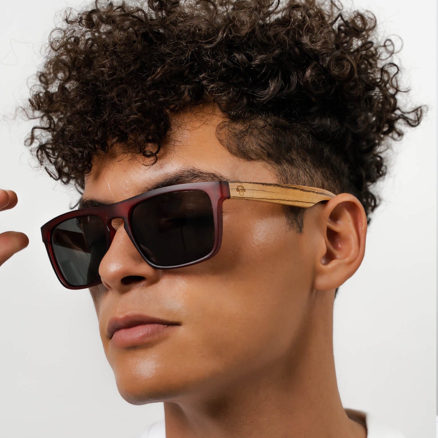 un modelo hombre de pelo rulo mirando en 45 grados hacia la camara con anteojos de sol rectangulares de color rojo con lentes oscuras y patas de madera natural #color-marco_wine