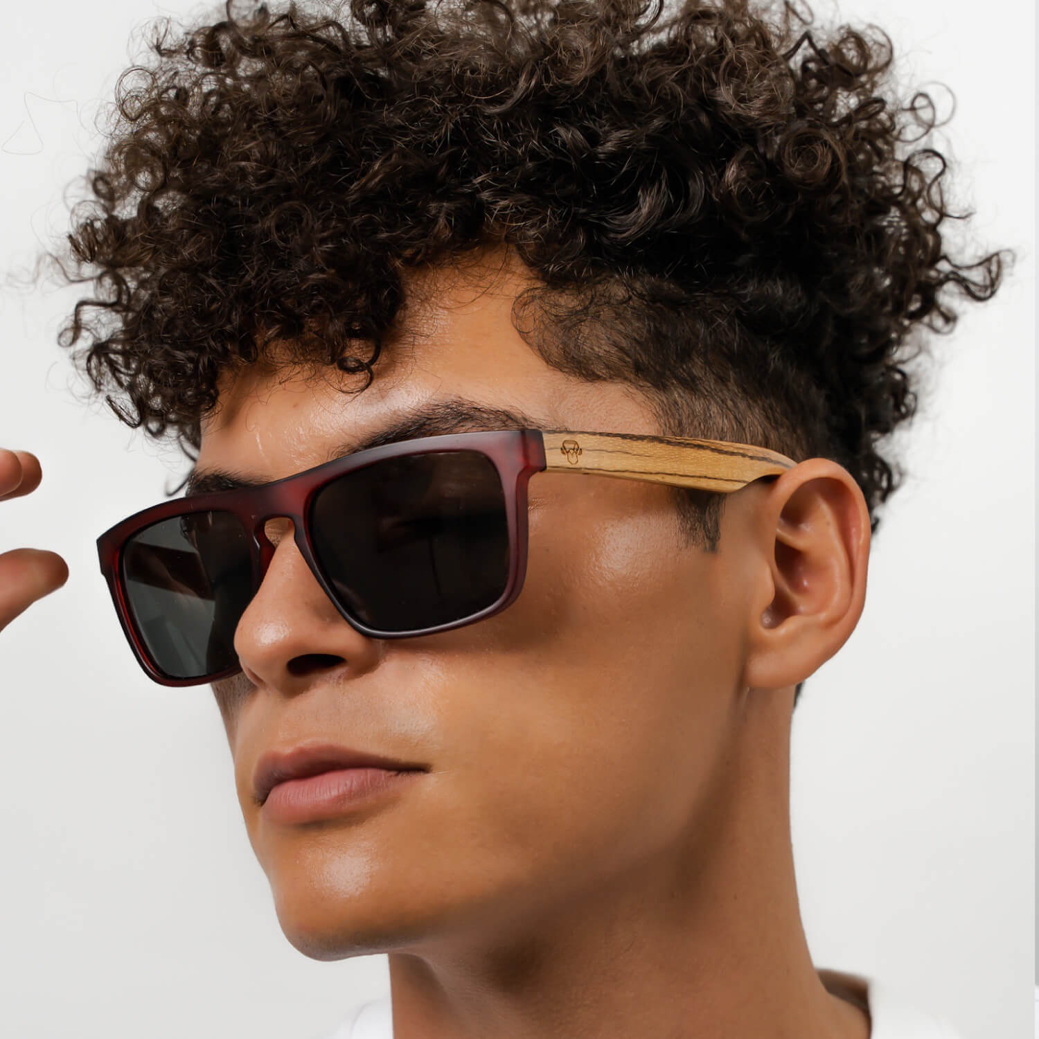 un modelo hombre de pelo rulo mirando en 45 grados hacia la camara con anteojos de sol rectangulares de color rojo con lentes oscuras y patas de madera natural #color-marco_wine