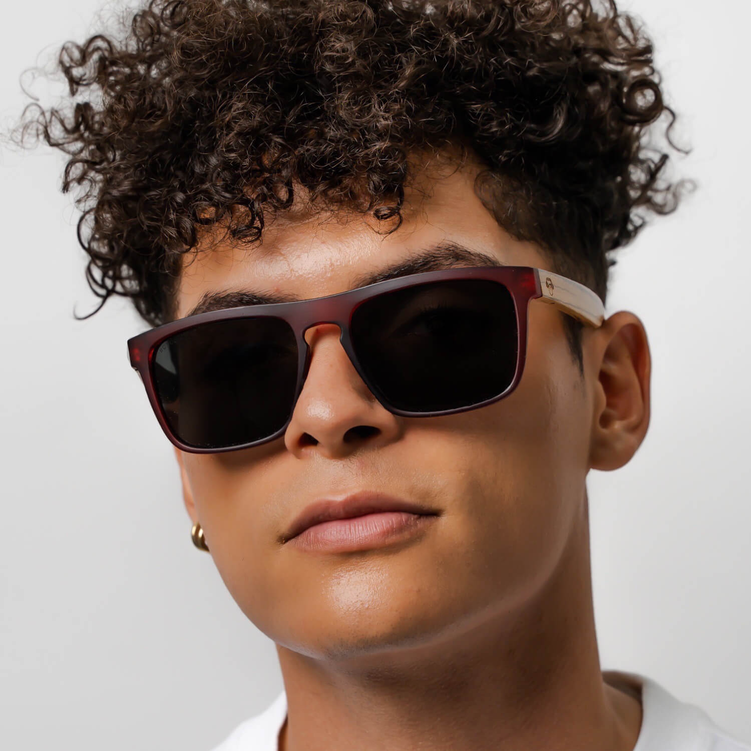 un modelo hombre de pelo rulo mirando en 25 grados hacia la camara con anteojos de sol rectangulares de color rojo con lentes oscuras y patas de madera natural #color-marco_wine