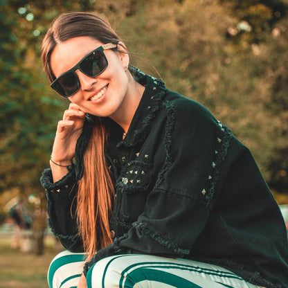 una mujer sonriendo mirando a la camara en 45 grados de pelo liso con chaqueta negra desgastada con tachas con anteojos de sol polarizados de color negro hecho con plastico reciclado y patas de madera natural #color-marco_negro