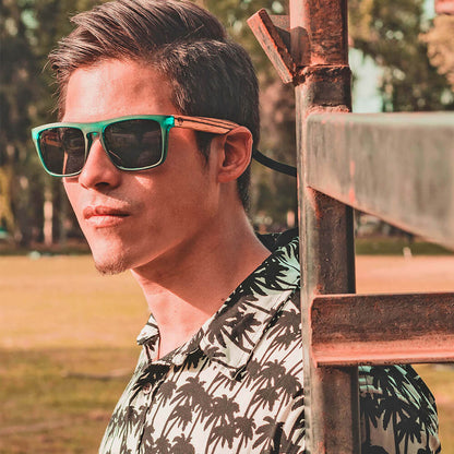un hombre serio en 45 grados con camisa blanca y un pattern de palmeras negras usando anteojos de sol polarizados de color verde hecho con plastico reciclado y patas de madera natural #color-marco_forest