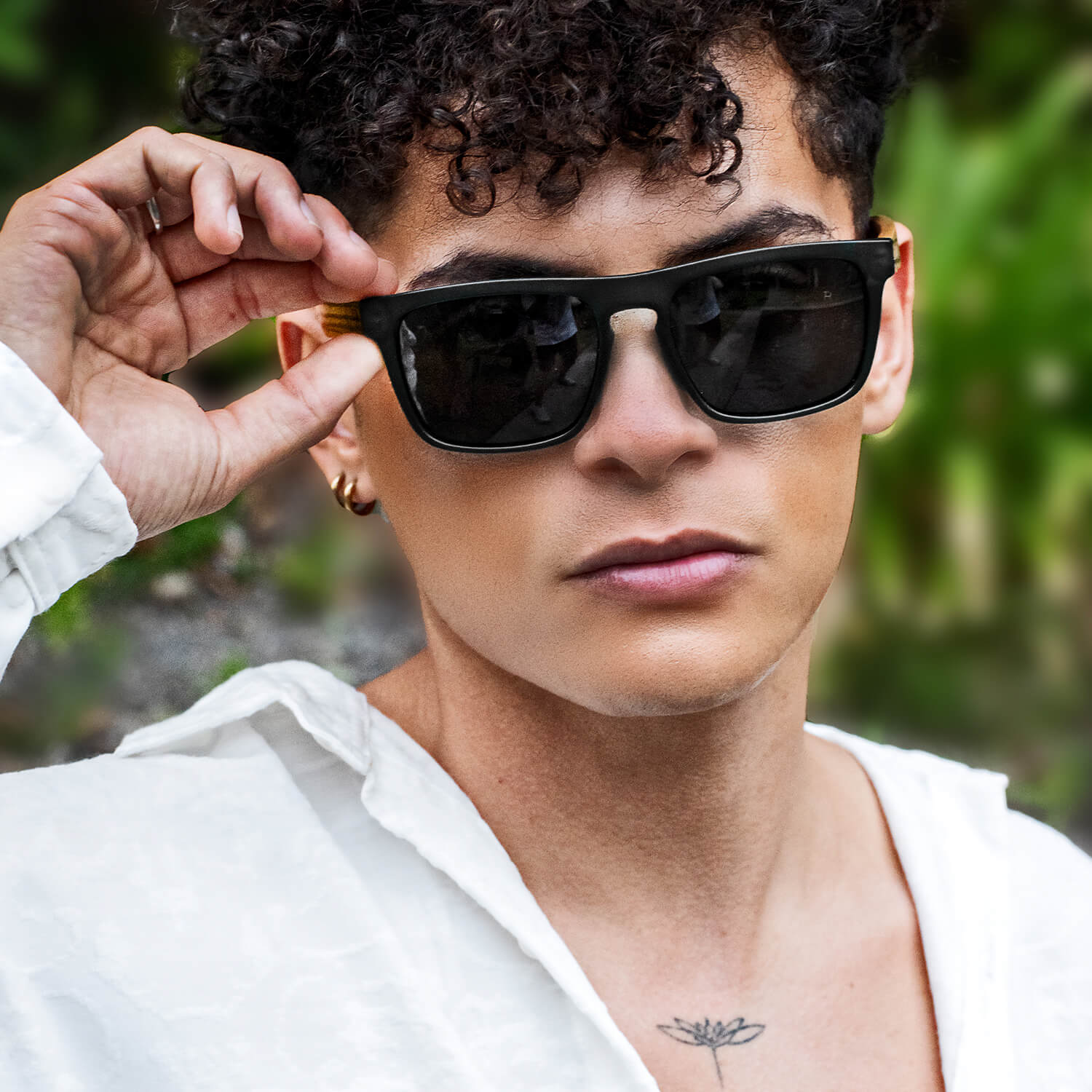 un modelo hombre de pelo rulo mirando en 25 grados hacia la camara con anteojos de sol rectangulares de color negro con lentes oscuras y patas de madera natural #color-marco_negro